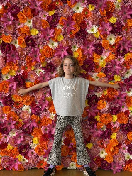A young child stands centered in front of a vibrant, colorful flower wall, posing for the camera